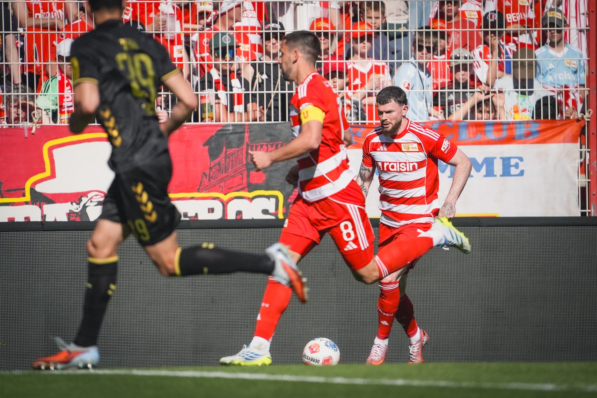 Spielszene im Fußball mit Spielern in roten und schwarzen Trikots. Zuschauer im Hintergrund mit Fanschals.