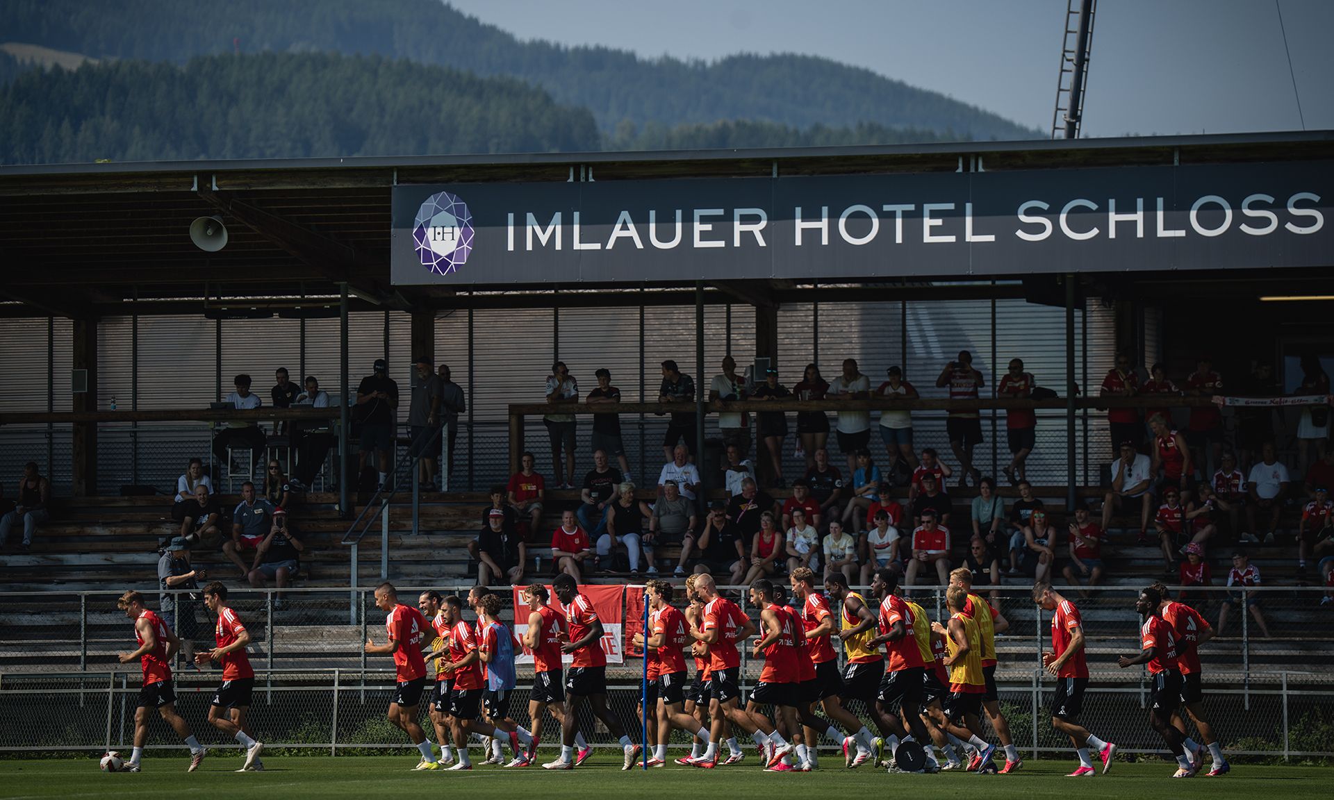 Fußballspieler in roten Trikots trainieren auf einem Platz, Zuschauer sitzen auf Tribünen im Hintergrund.酒店标志在其上方。