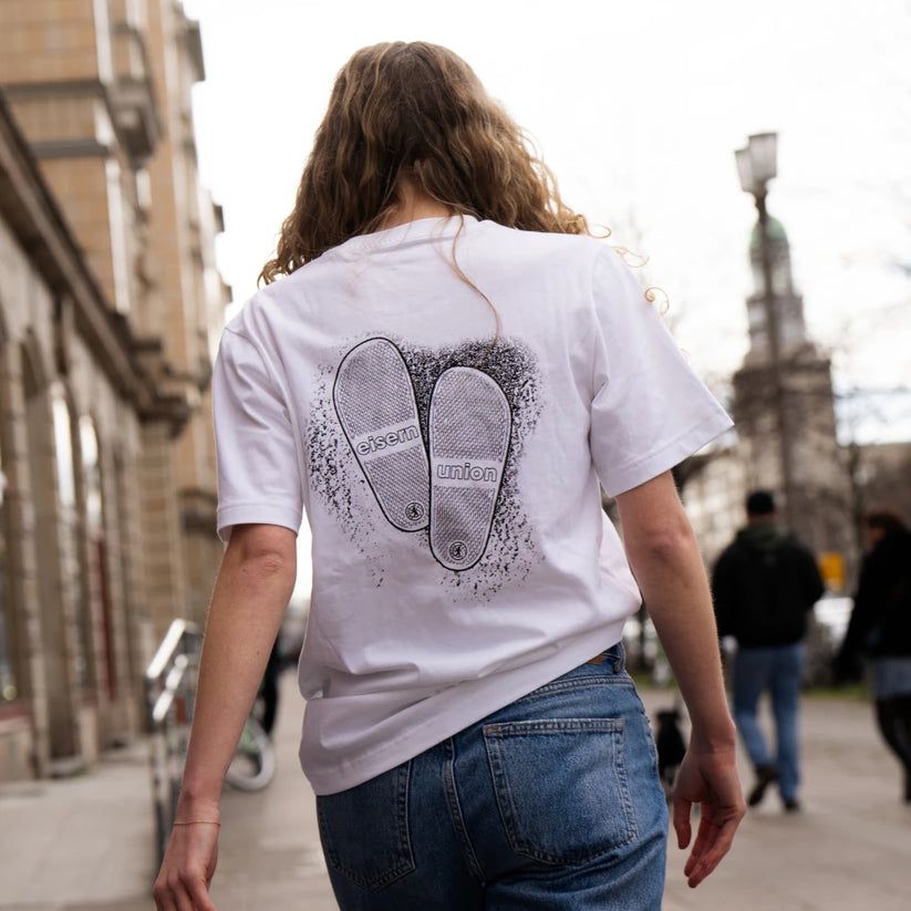 Eine Person trägt ein weißes T-Shirt mit einem Druck von Schuhabdrücken auf dem Rücken, während sie auf einer Straße geht.