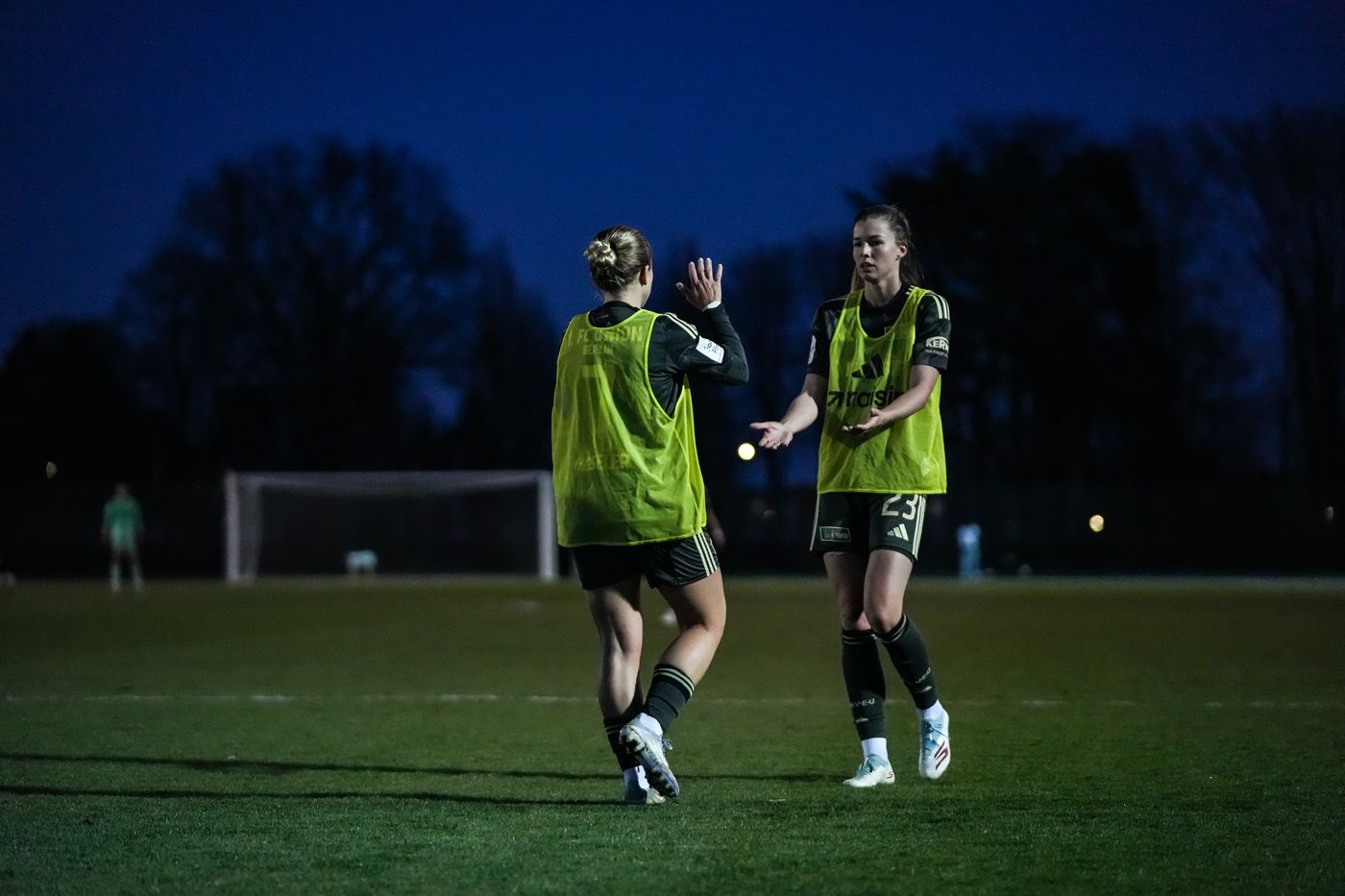 Zwei Fußballspielerinnen in gelben Trainingswesten interagieren auf einem rasierten Spielfeld bei Nacht.