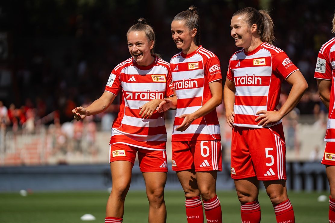 Drei Fußballspielerinnen in rot-weißen Trikots stehen lachend zusammen auf dem Feld.