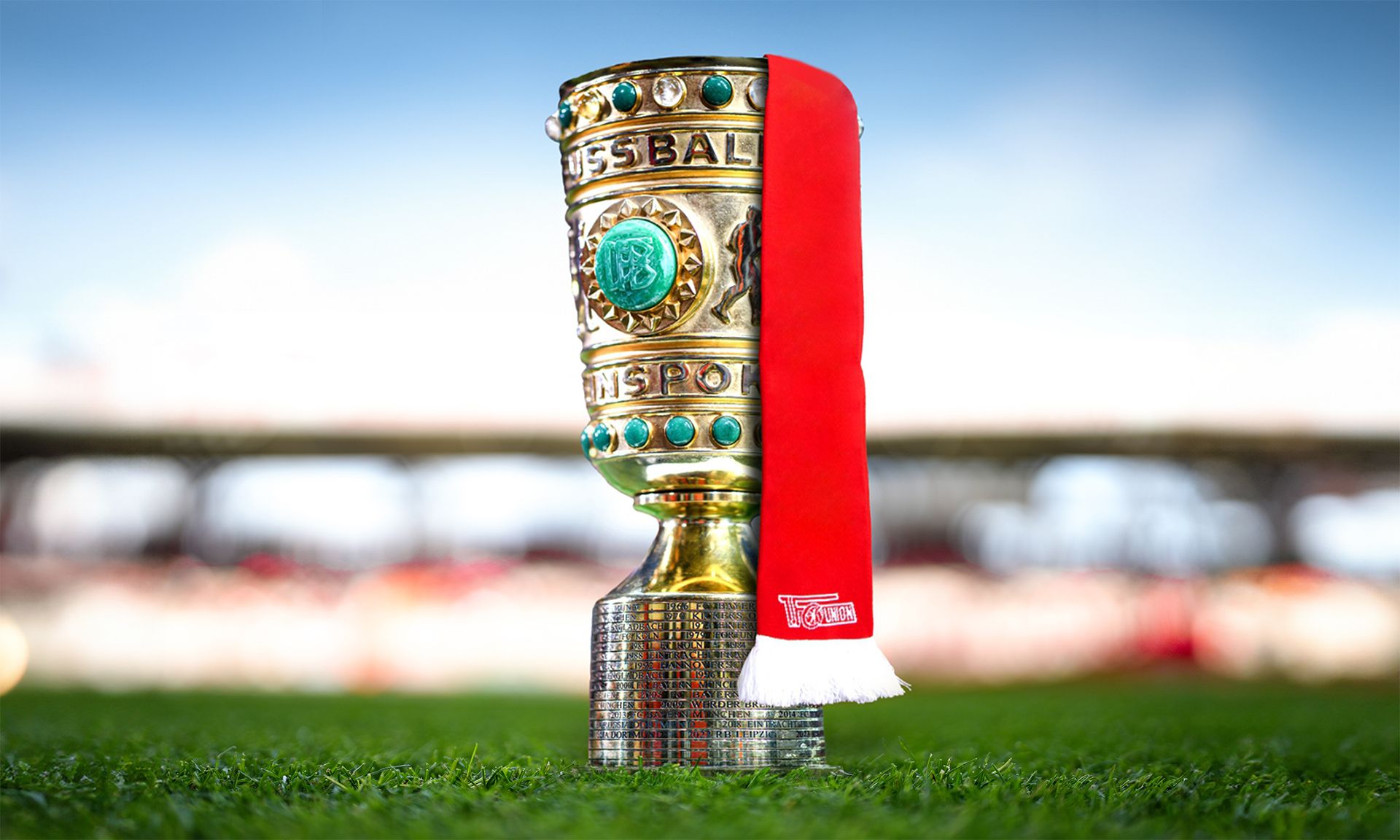 Ein Fußballpokal mit grünen Edelsteinen und rotem Schal steht auf einem grünen Rasen.