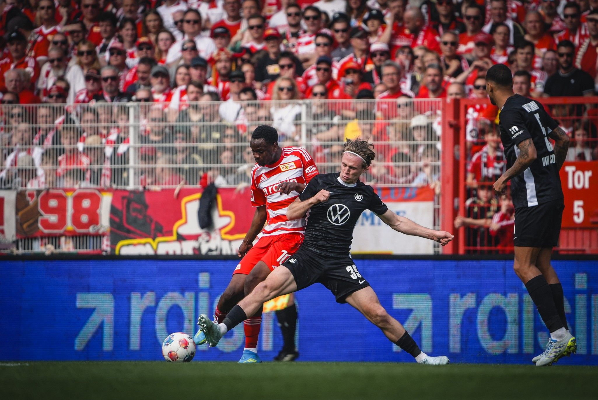 Ein Fußballspiel mit einem Spieler in rotem Trikot, der einen anderen Spieler in schwarzem Trikot anvisiert, im Hintergrund jubelnde Fans.