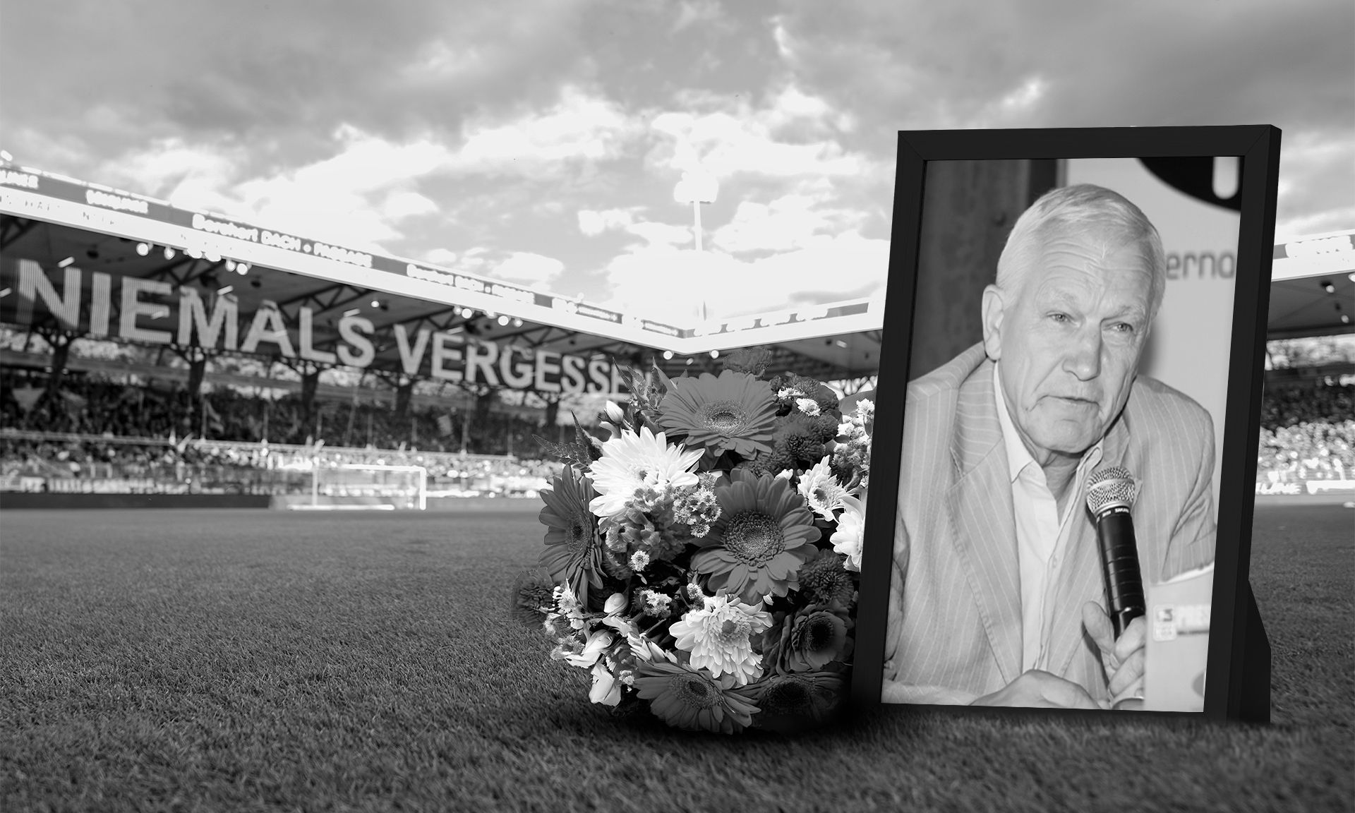 Ein Grabstein mit einem Portrait eines älteren Mannes, flankiert von einem Blumenarrangement, vor einem Fußballstadion.