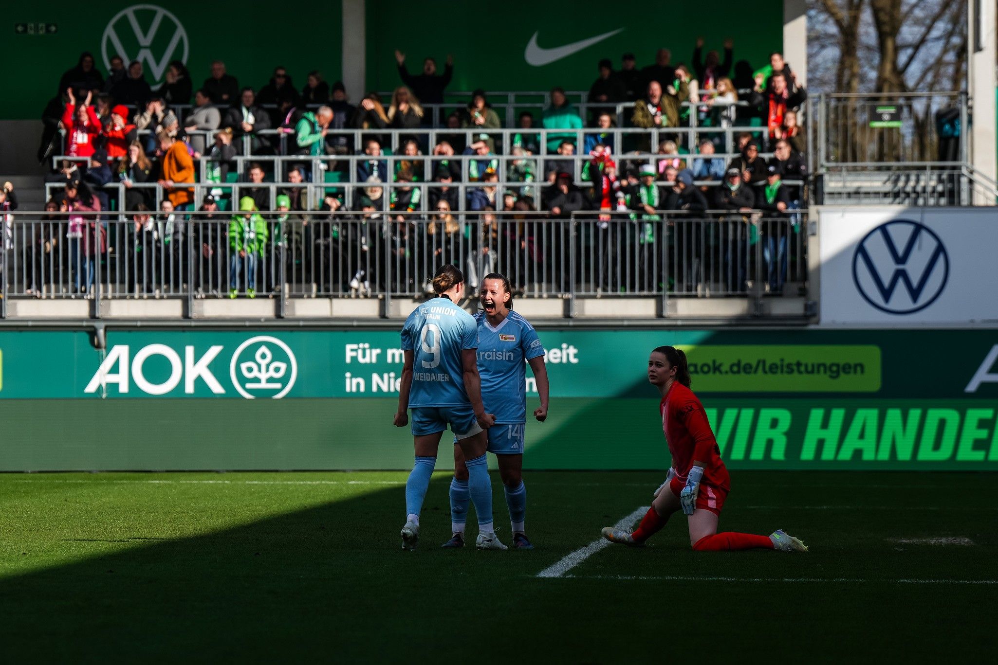 Zwei Fußballspieler feiern ein Tor, während ein Torwart auf dem Boden sitzt. Zuschauer im Hintergrund beobachten das Geschehen.