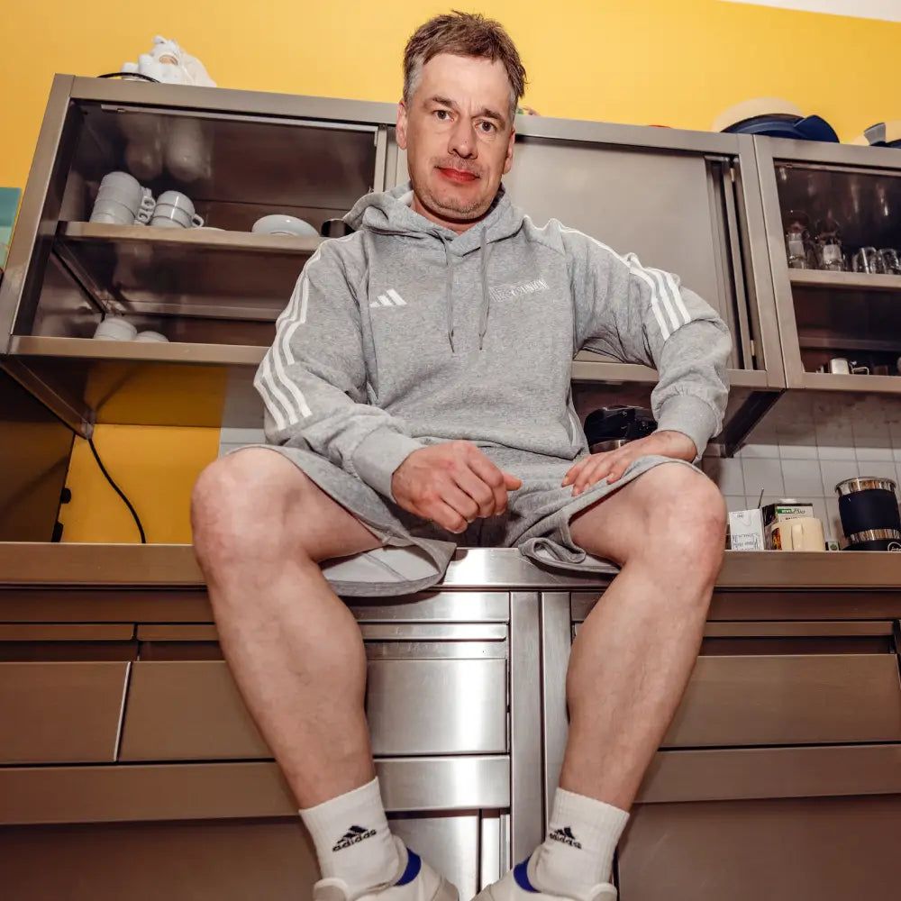 Mann in grauem Jogginganzug sitzt auf einer Küchenarbeitsplatte, umgeben von Küchenschränken und Geräten.