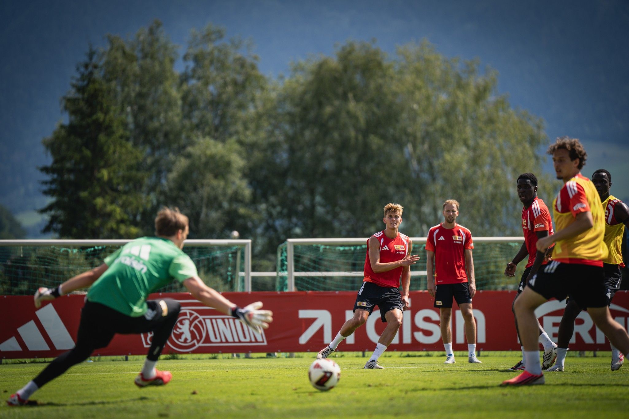 Fußballtraining auf dem Rasen mit Spielern in roten Trikots, einem Torwart in grün und einem Ball. Hintergrund mit Bäumen und Bergen.