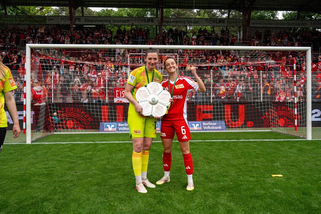 Zwei Fußballspielerinnen posieren mit einer Trophäe vor einer jubelnden Fangemeinde im Hintergrund.