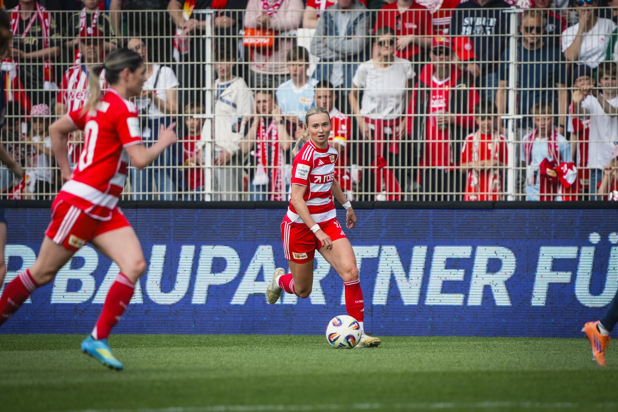 Zwei Frauenfußballspielerinnen in roten Trikots während eines Spiels, im Hintergrund jubelnde Fans.