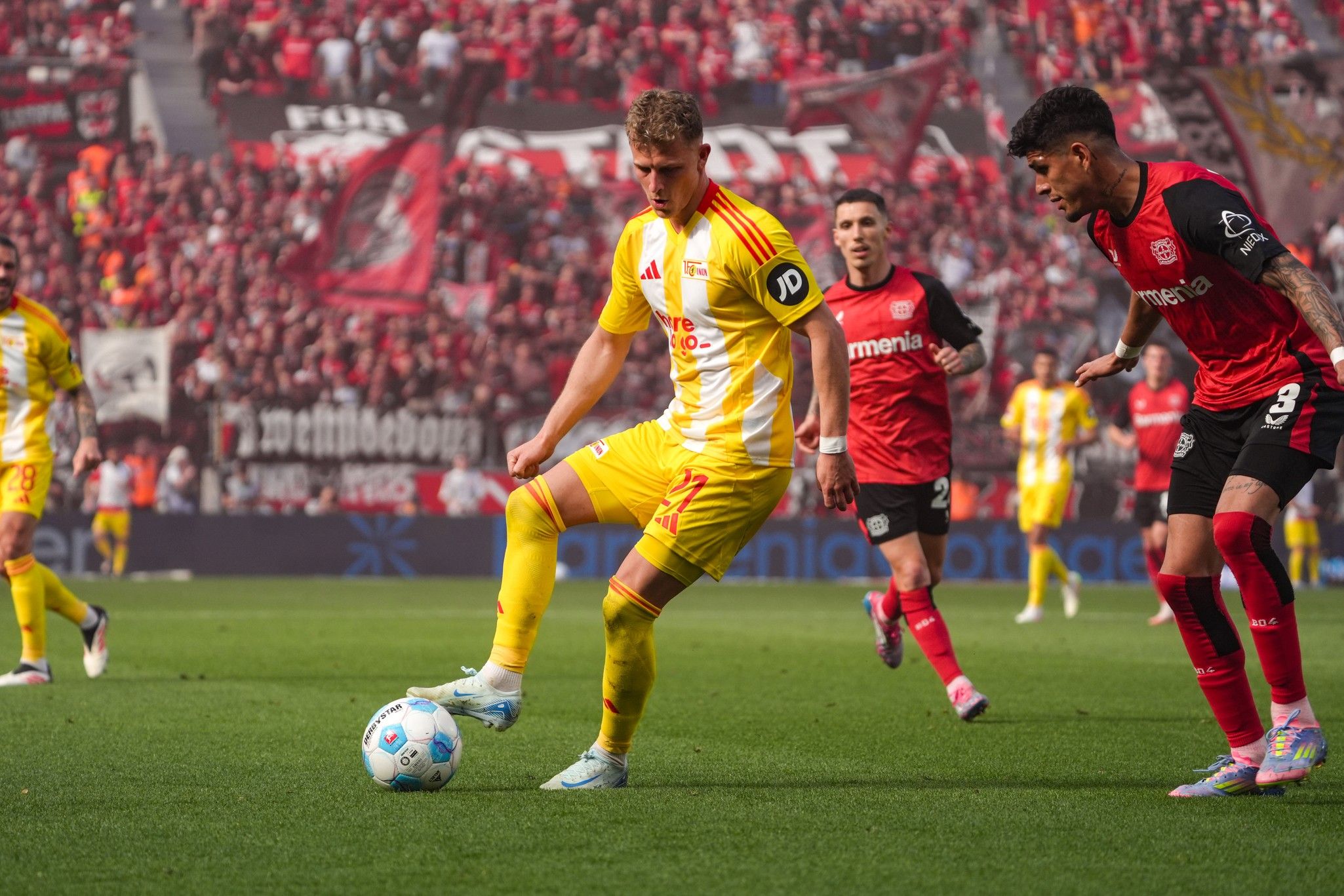 Fußballspiel mit Spielern in gelben und roten Trikots. Ein Spieler passt den Ball auf einem grünen Spielfeld. Fans im Hintergrund.