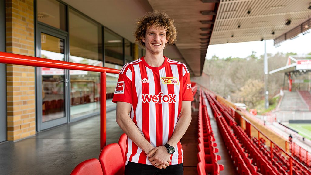 A smiling man in a red-and-white striped soccer jersey stands in a stadium with empty seats.