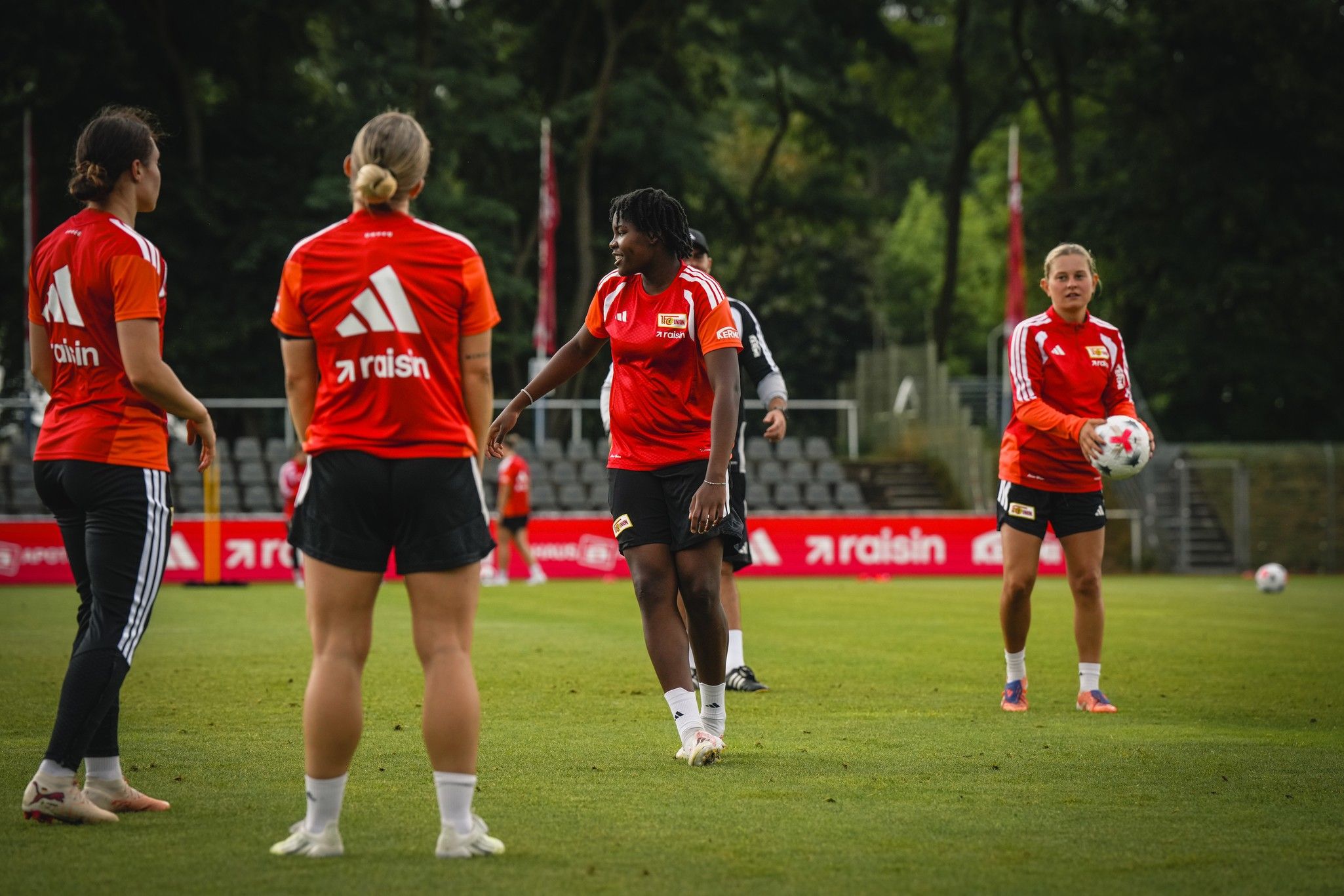 Frauenfußballmannschaft beim Training auf dem Platz, Spielerinnen in roten Trikots diskutieren und üben mit einem Ball.