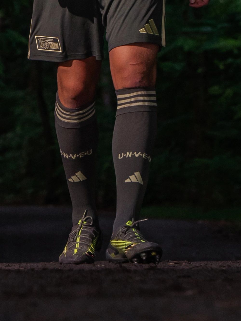 Beine einer Person in Fußballkleidung mit grauen Shorts, langen Socken und speziellen Fußballschuhen, auf einem dunklen Weg.