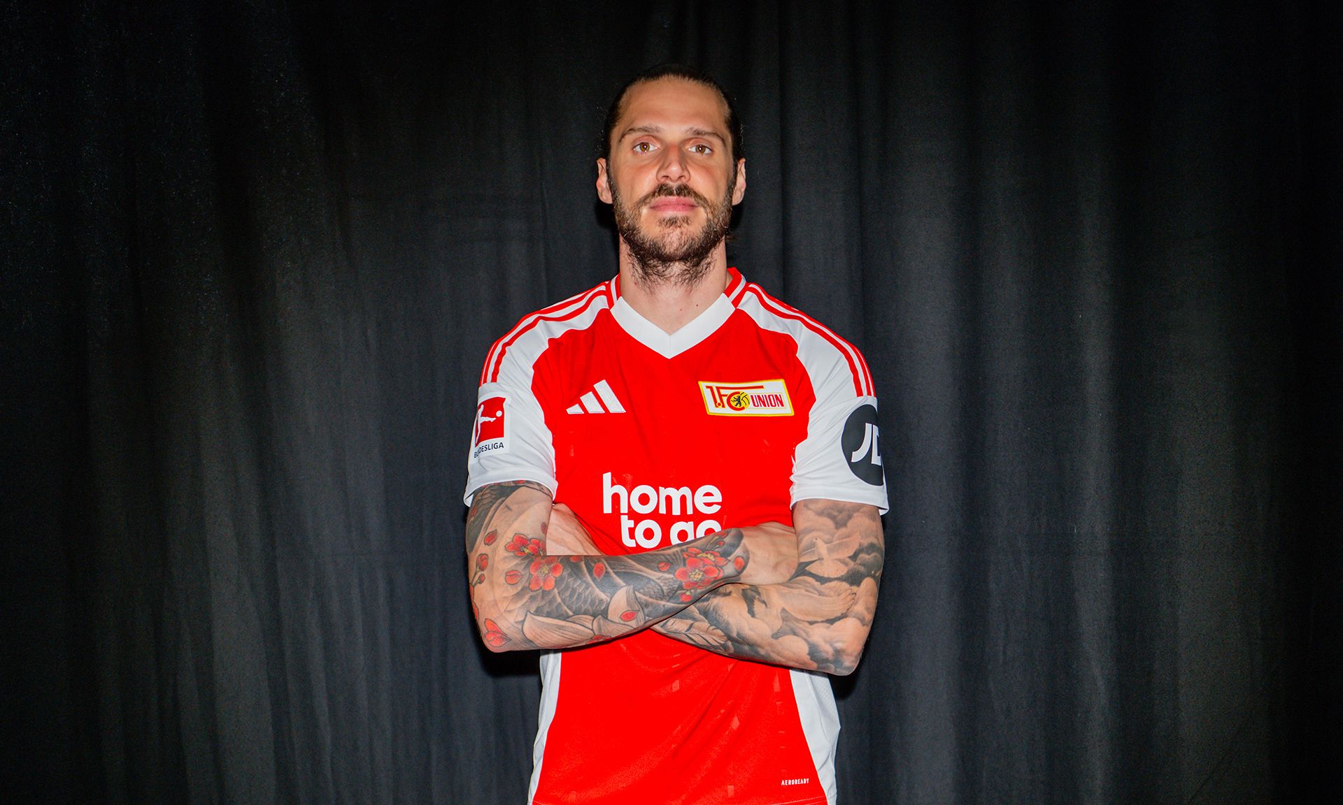 Ein sportlicher Mann mit tätowierten Armen in einem roten Fußballtrikot mit weißen Akzenten steht mit verschränkten Armen vor schwarzem Hintergrund.