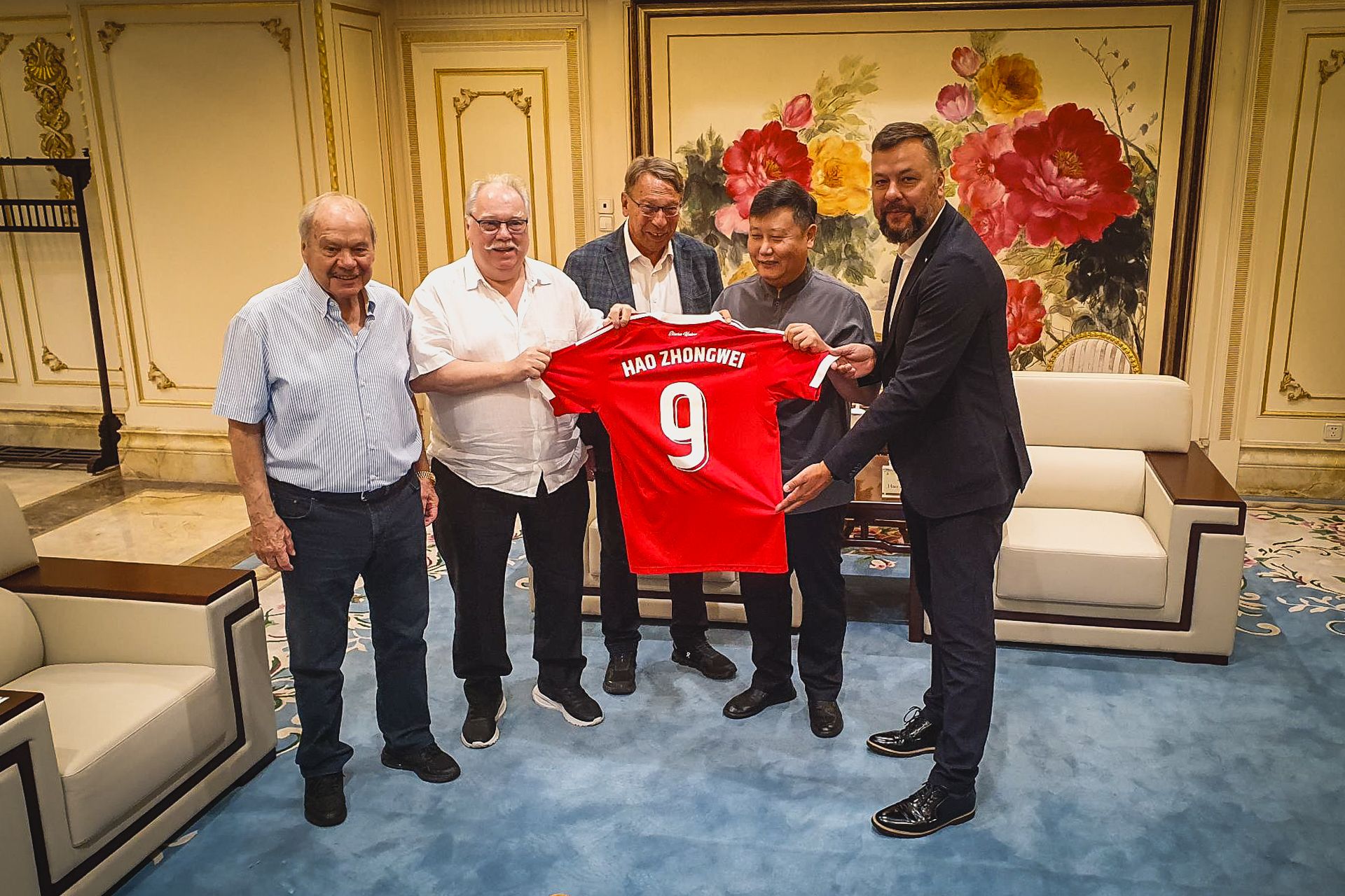 Gruppierung von fünf Personen in einem eleganten Raum, die ein rot gestreiftes Trikot mit der Aufschrift "Hao Zhongwei 9" halten.