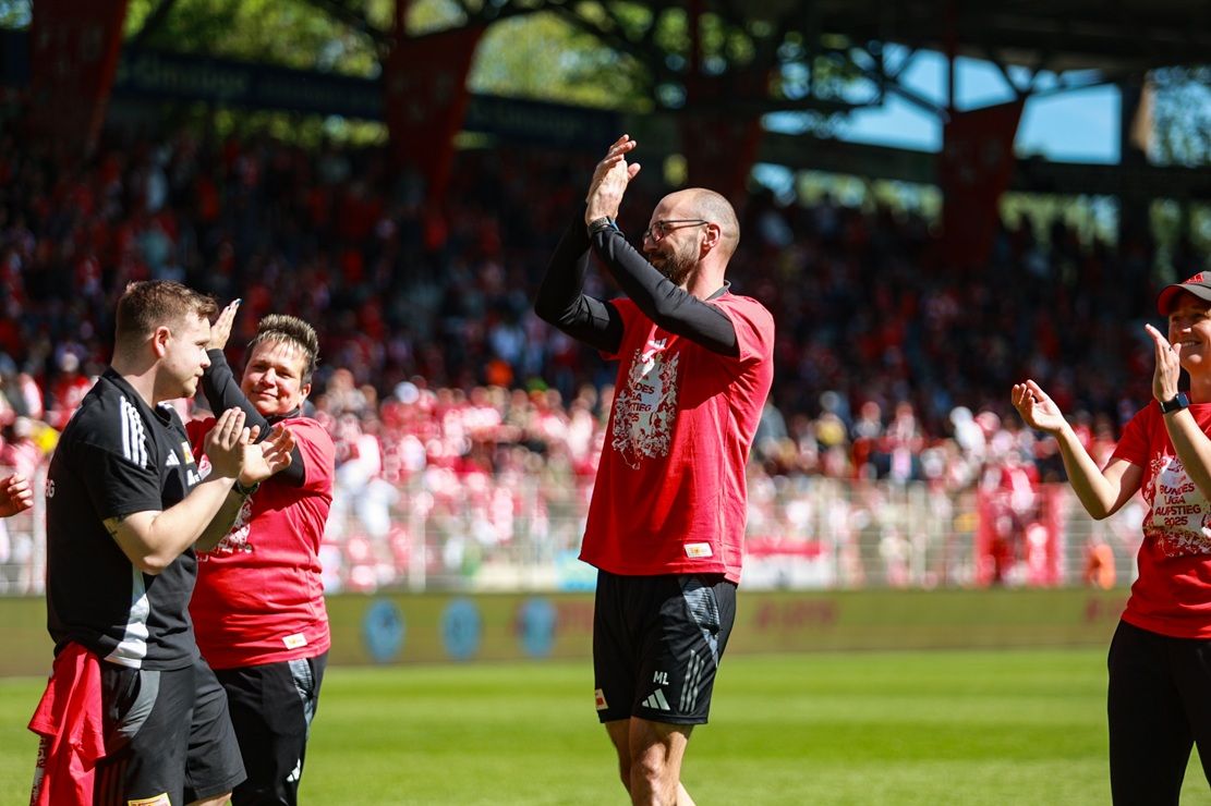 Personen in roten T-Shirts applaudieren auf einem Sportfeld vor einer jubelnden Menge.