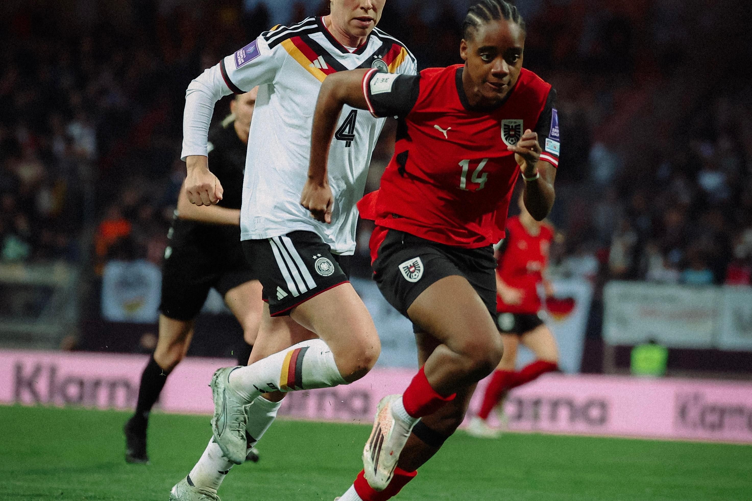 Zwei Fußballspielerinnen in Aktion auf dem Spielfeld, eine in rotem Trikot, die andere in weißem Trikot mit schwarzer Hose.