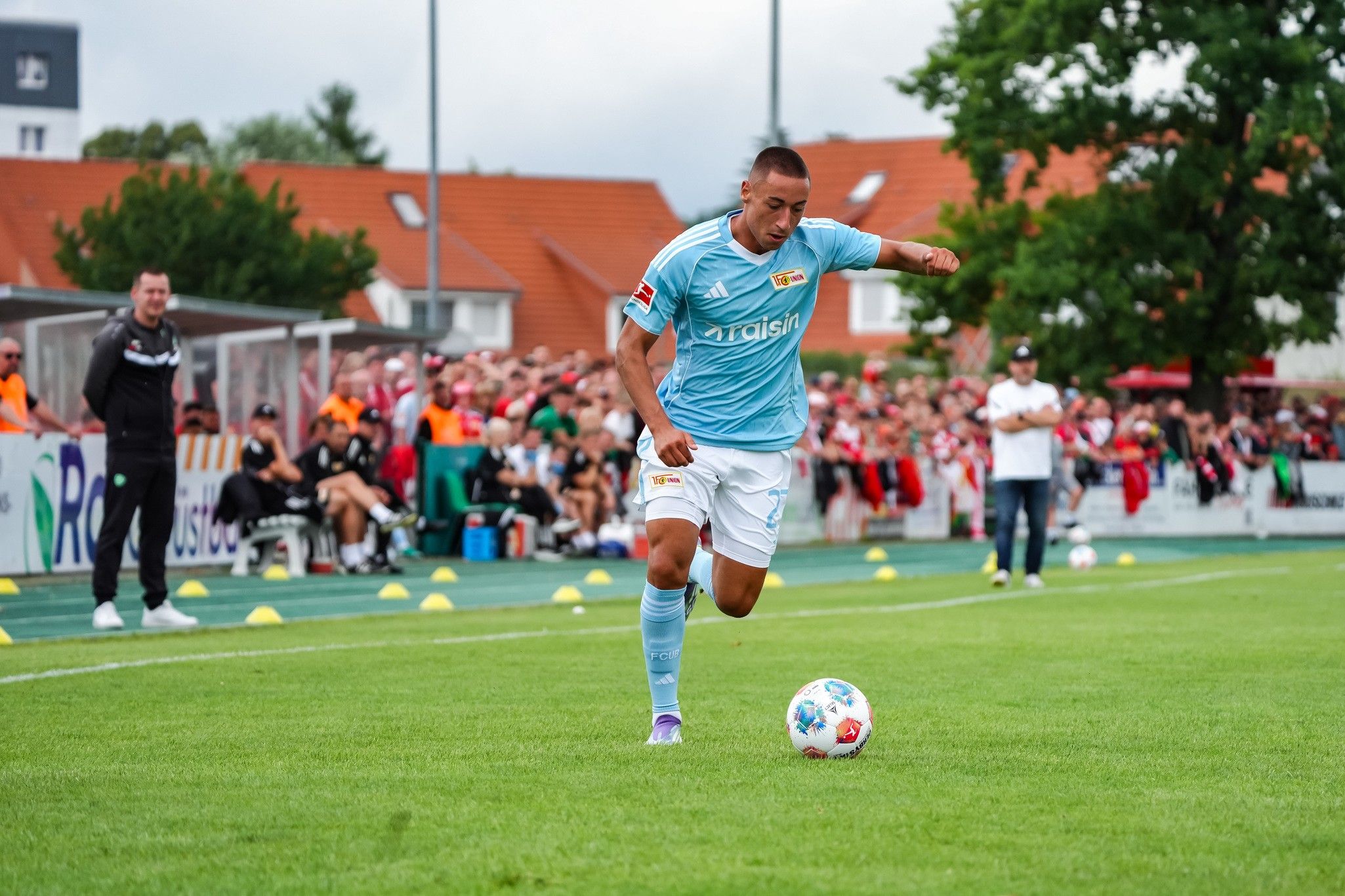Ein Fußballspieler in hellblauer Trikot führt einen Ball auf einem grünen Platz vor einer Menge zuschauender Fans.