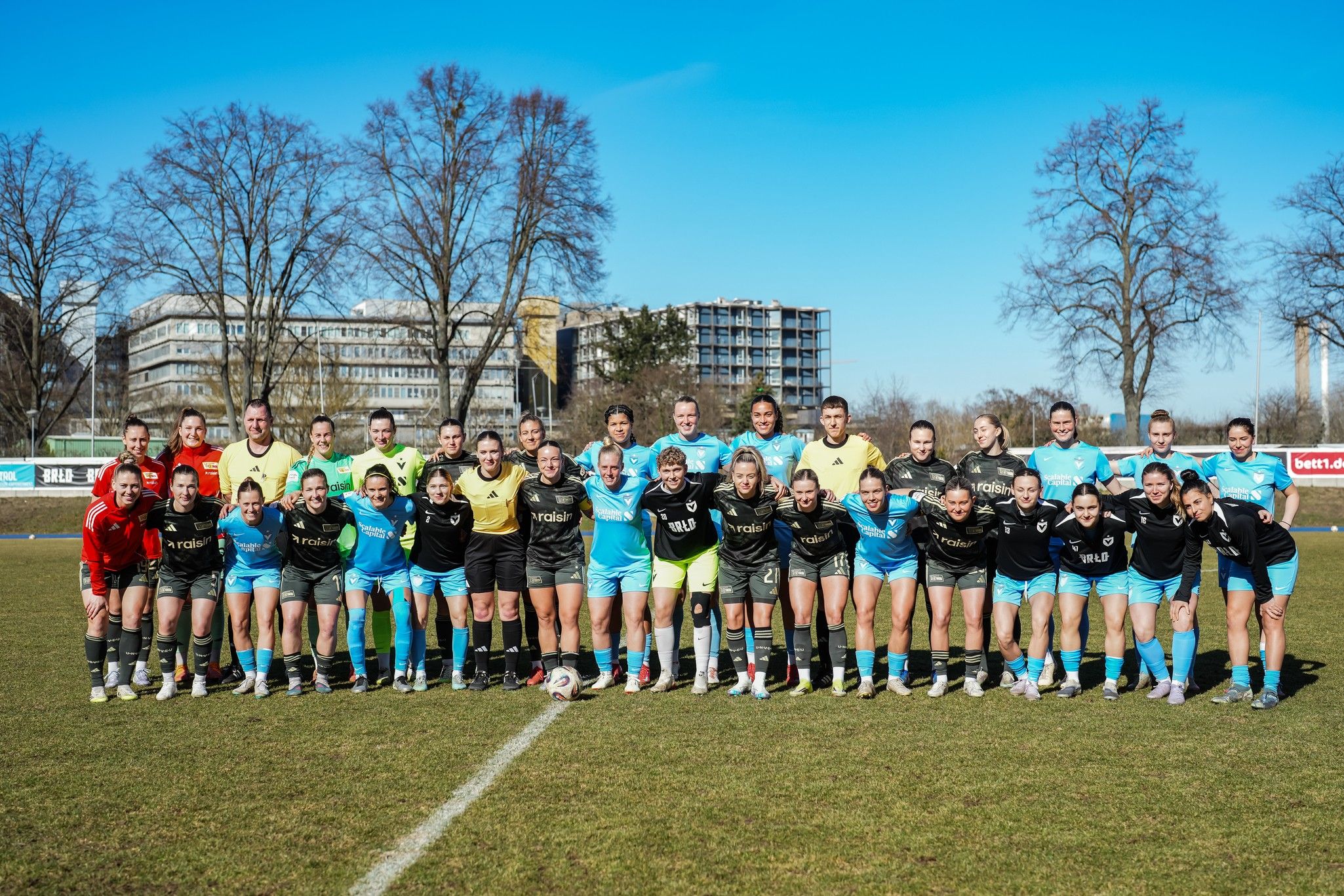 Gruppenfoto von zwei Frauenfußballmannschaften auf einem Sportplatz, im Hintergrund Bäume und Gebäude, klarer Himmel.