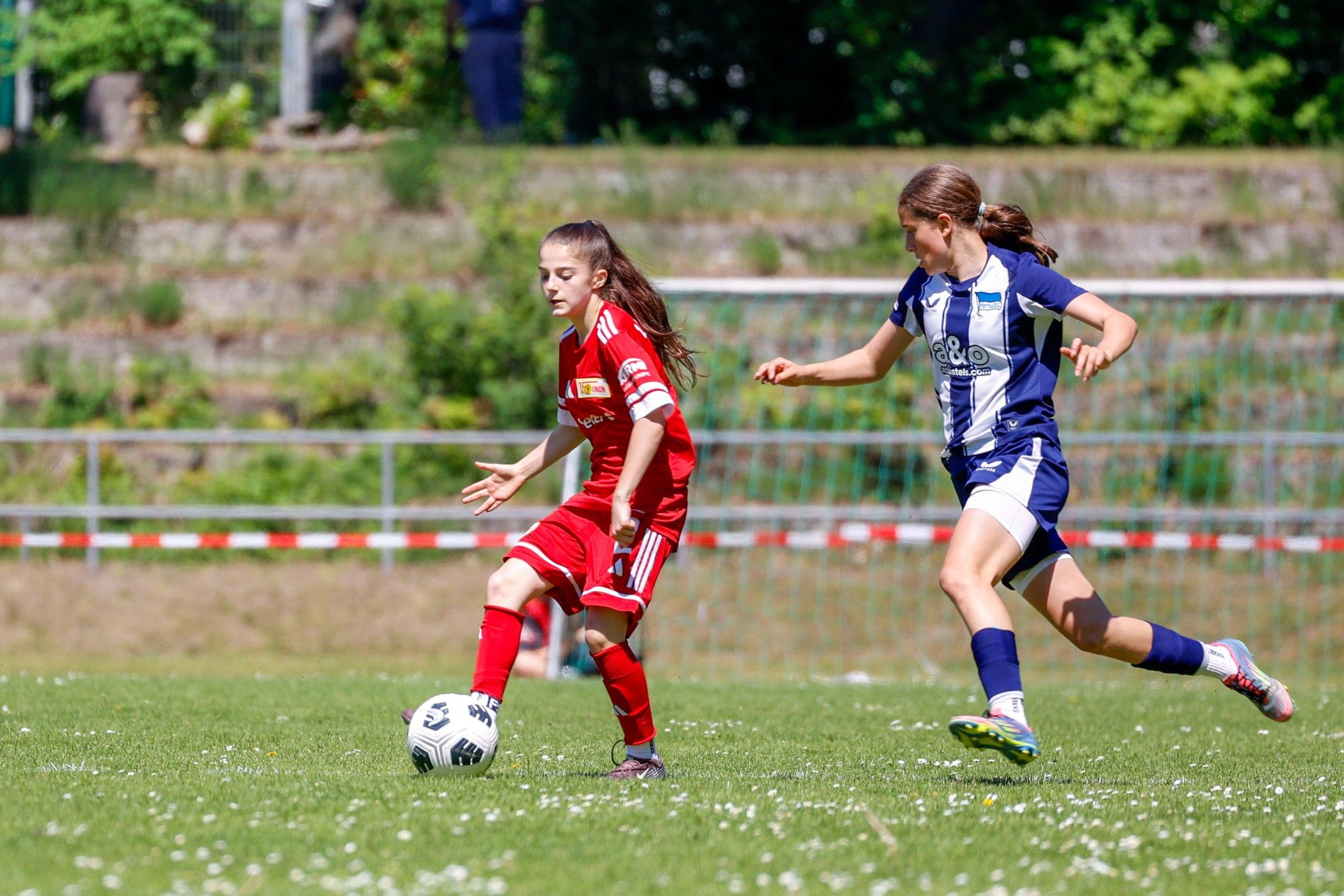 Zwei Mädchen spielen Fußball auf einem grünen Platz; eine trägt ein rotes, die andere ein blau-weißes Trikot.