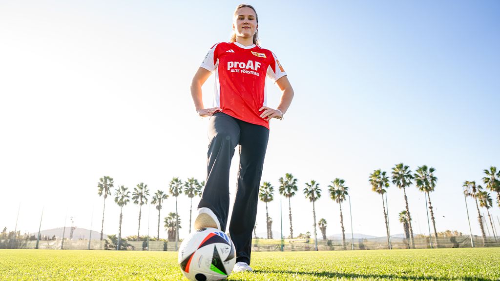 Eine sportliche Frau steht auf einem Fußballfeld, trägt ein rotes Trikot und hat einen Fußball vor sich auf dem Rasen.