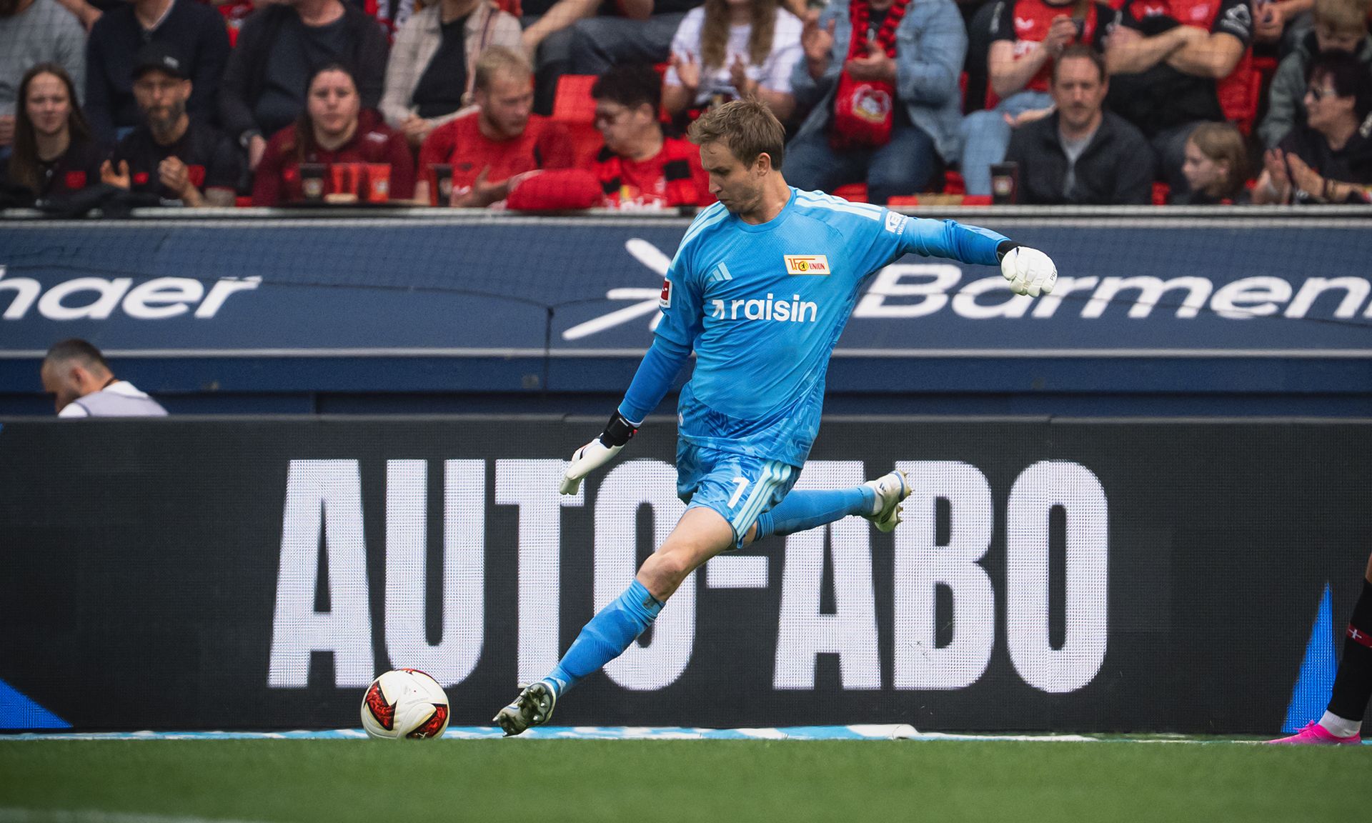 Torwart in blauer Ausrüstung führt einen Abschlag während eines Fußballspiels aus, Zuschauer im Hintergrund.