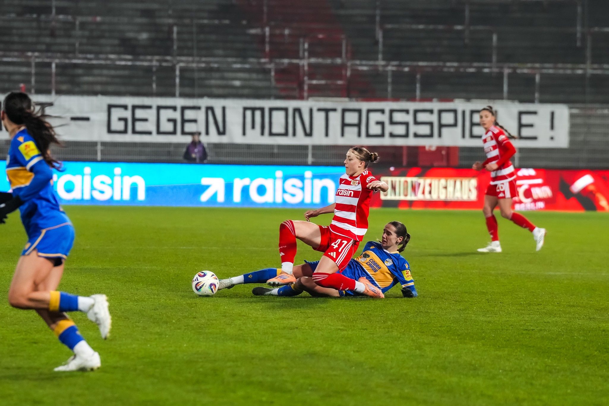 Eine Fußballspielszene mit zwei Spielerinnen, die im Kampf um den Ball am Boden liegen. Im Hintergrund ein Banner.