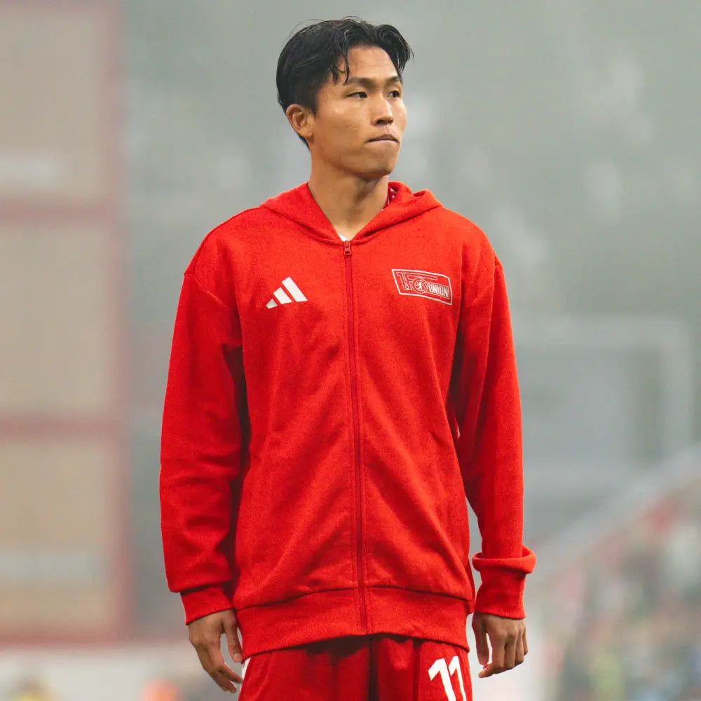 Ein Fußballspieler in roter Trainingskleidung mit einer Kapuze steht auf dem Platz und schaut konzentriert.