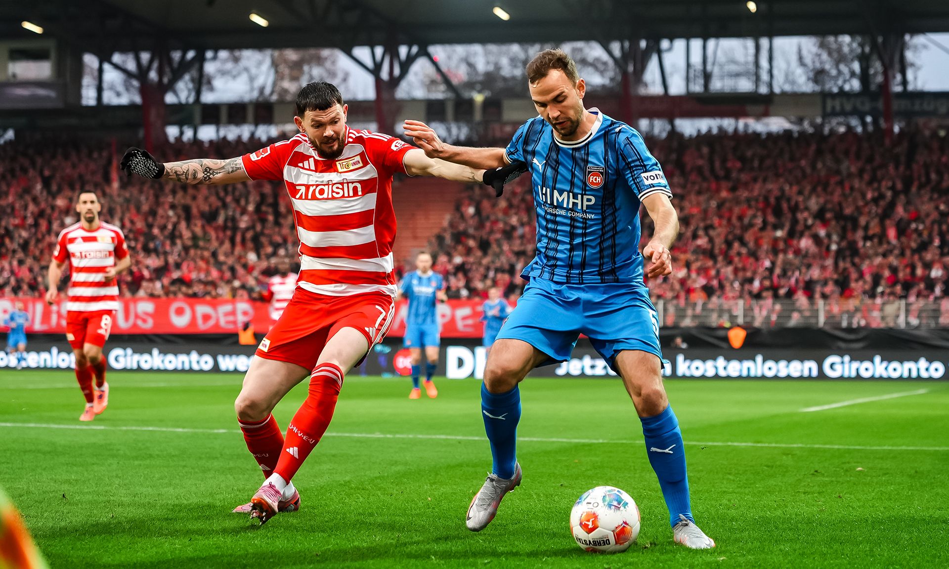 Zwei Fußballspieler kämpfen um den Ball auf dem Spielfeld während eines Spiels, im Hintergrund ist die Zuschauertribüne zu sehen.