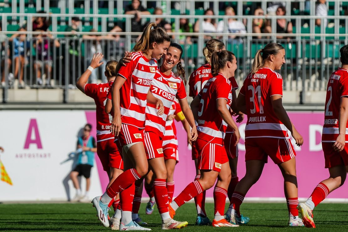 Mehrere Fußballspielerinnen in roten Trikots feiern ein Tor auf einem Stadionfeld. Zuschauer im Hintergrund.