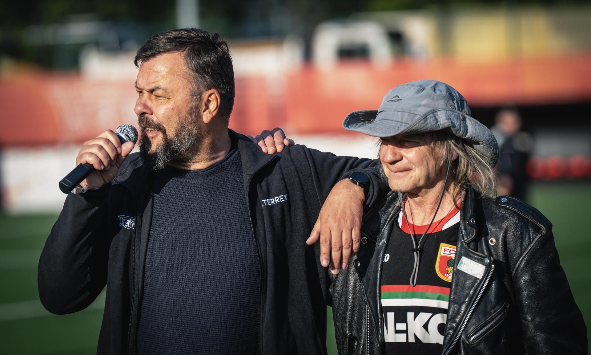 Zwei Männer stehen auf einem Sportplatz, einer spricht ins Mikrofon, während der andere ihm lächelnd die Schulter berührt.