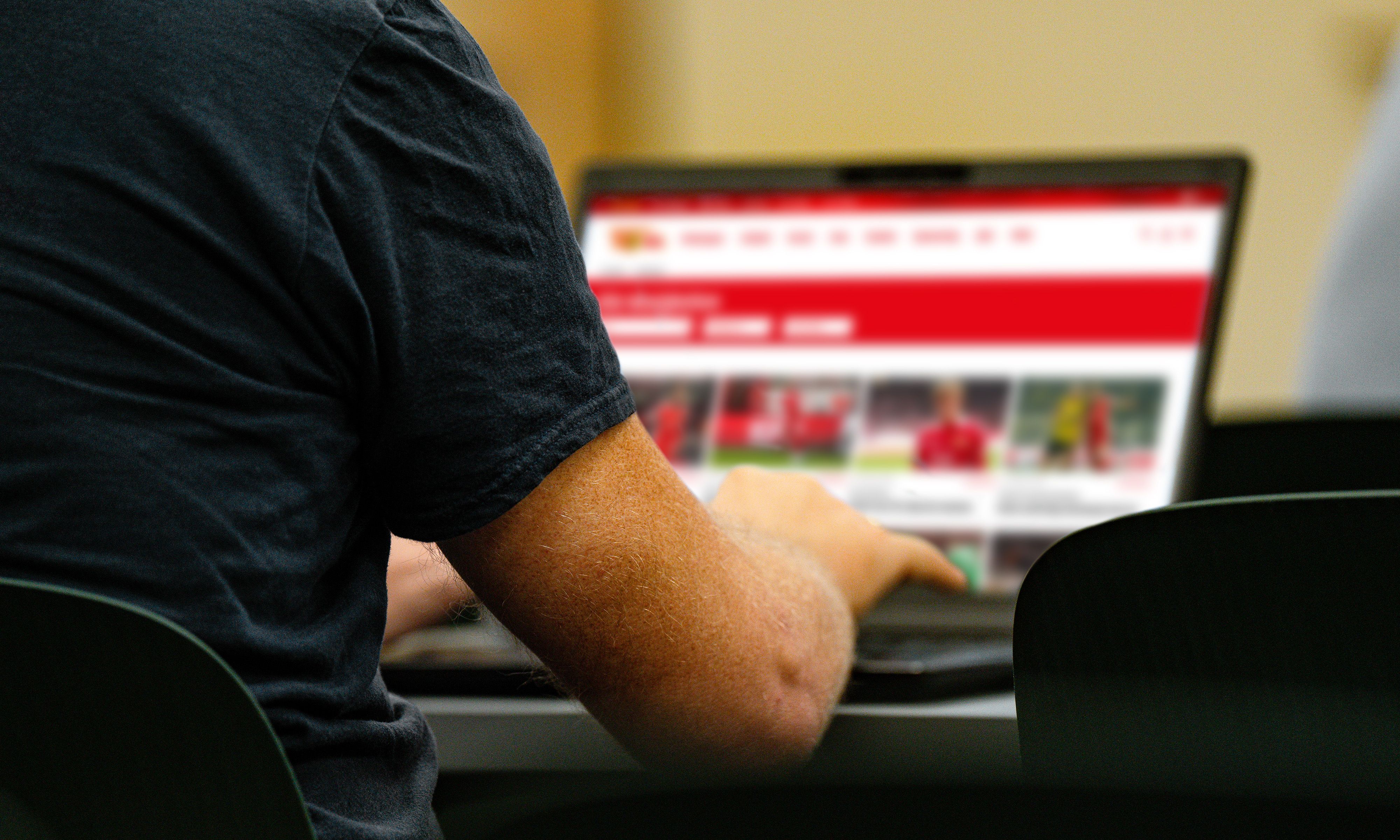 Eine Person sitzt an einem Tisch und benutzt einen Laptop mit einem geöffneten Bildschirm, auf dem eine Webseite angezeigt wird.