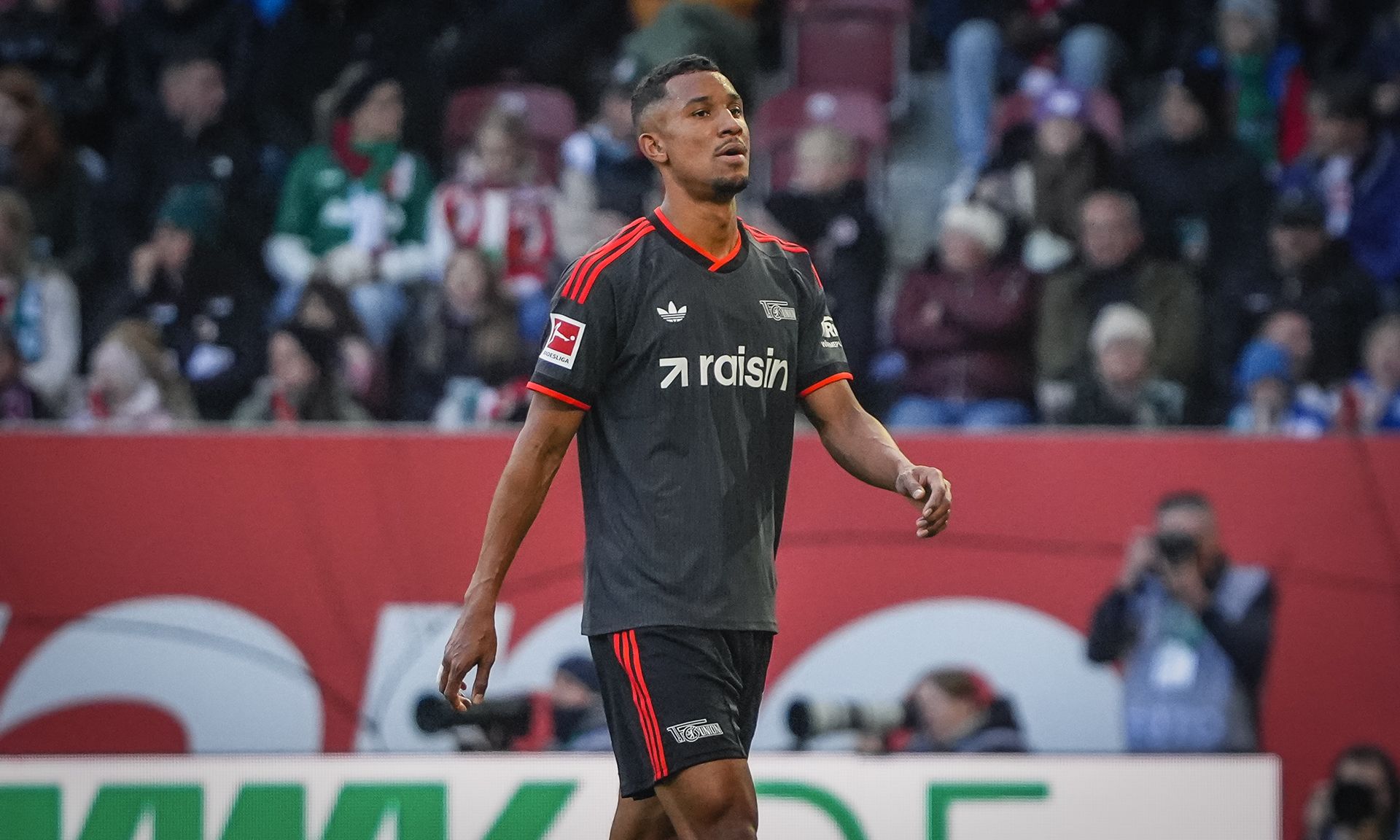 Ein Fußballspieler in grauem Trikot von hinten auf dem Spielfeld, umgeben von jubelnden Fans.