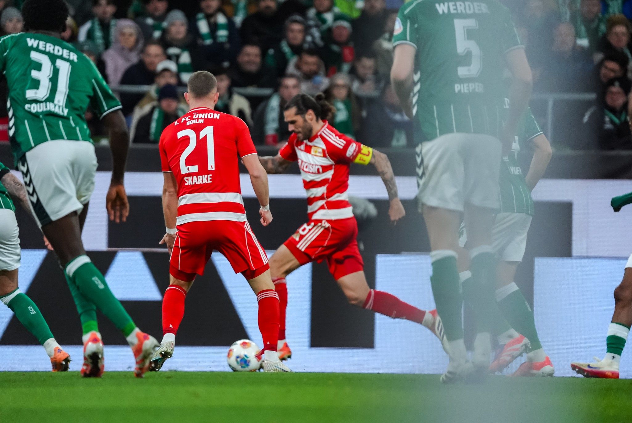 Zwei Fußballspieler in roten Trikots sind im Spiel, während grün gekleidete Gegner in der Nähe sind. Action auf dem Rasen.