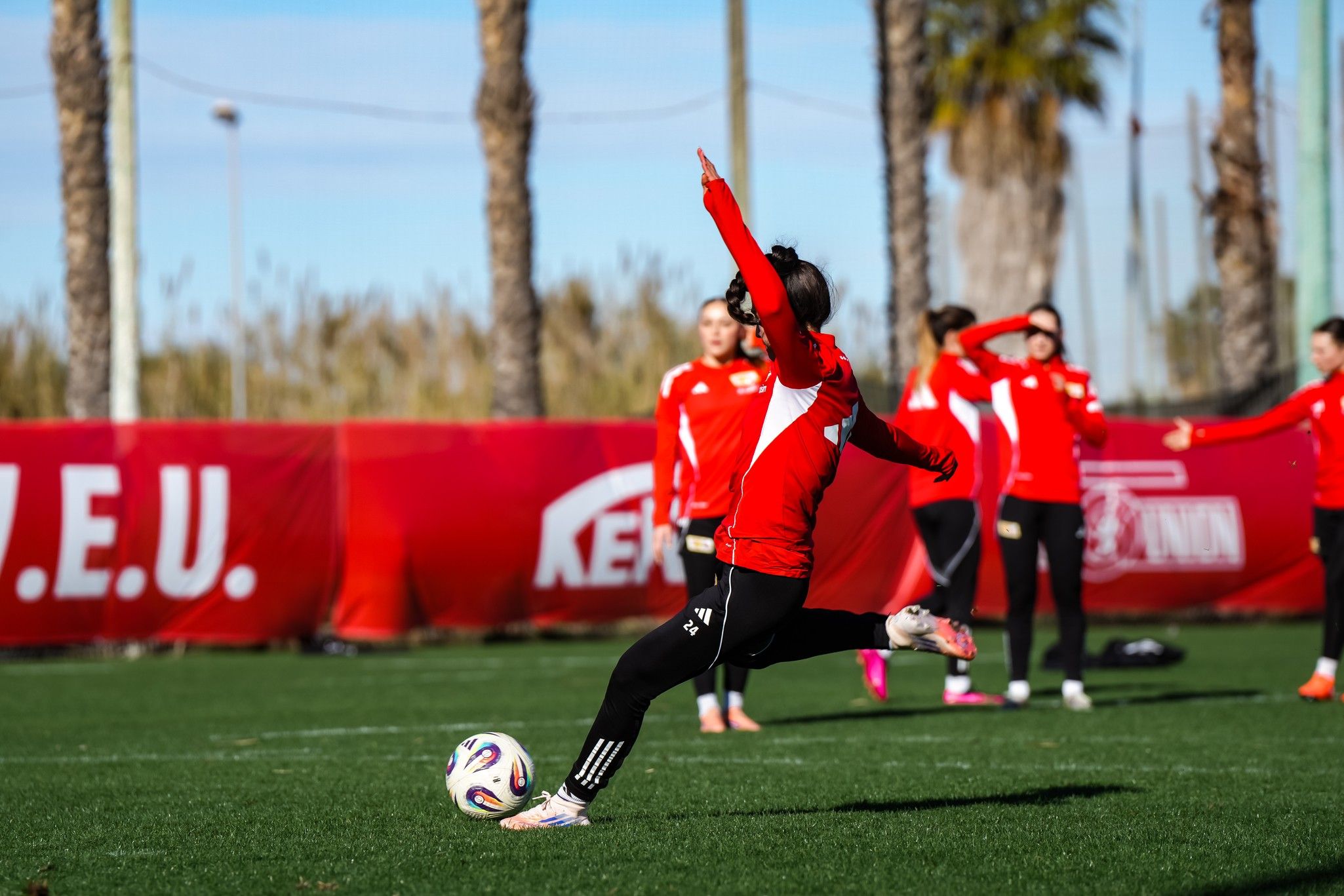 Eine weibliche Spielerin tritt den Ball während eines Fußballtrainings auf einem grünen Platz mit Palmen im Hintergrund.