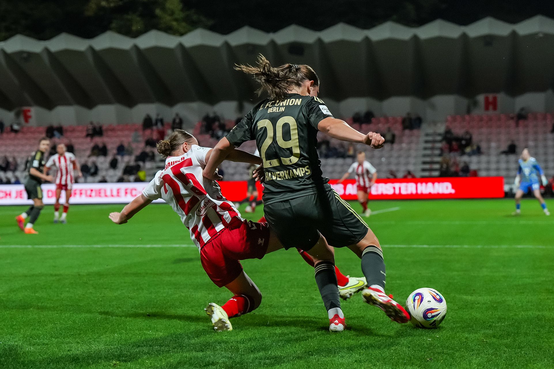 Fußballspiel mit zwei Spielern im Zweikampf auf dem Spielfeld, Zuschauer im Hintergrund.