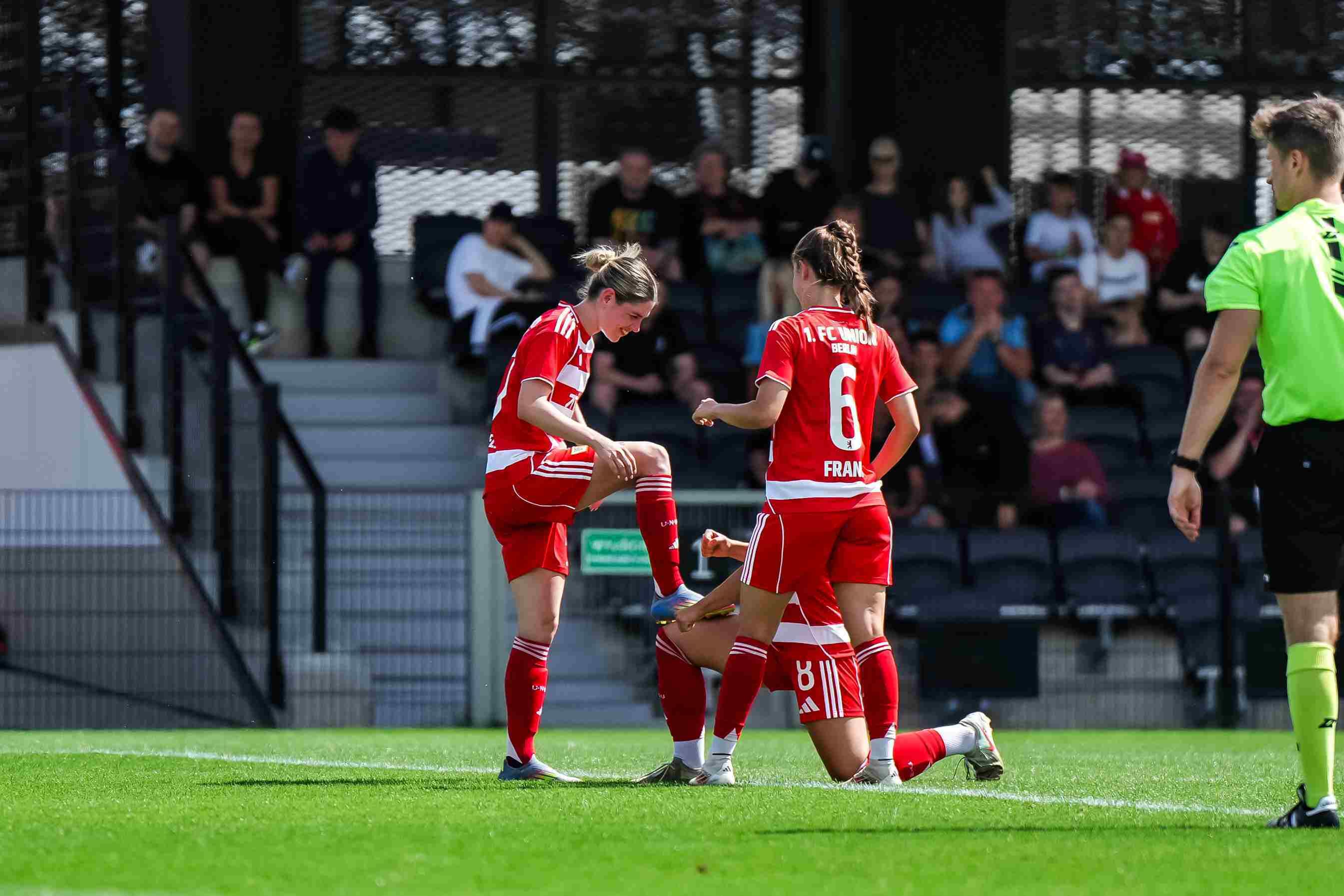 Drei Fußballspielerinnen in roten Trikots feiern auf dem Spielfeld, während ein Schiedsrichter in Gelb in der Nähe steht.