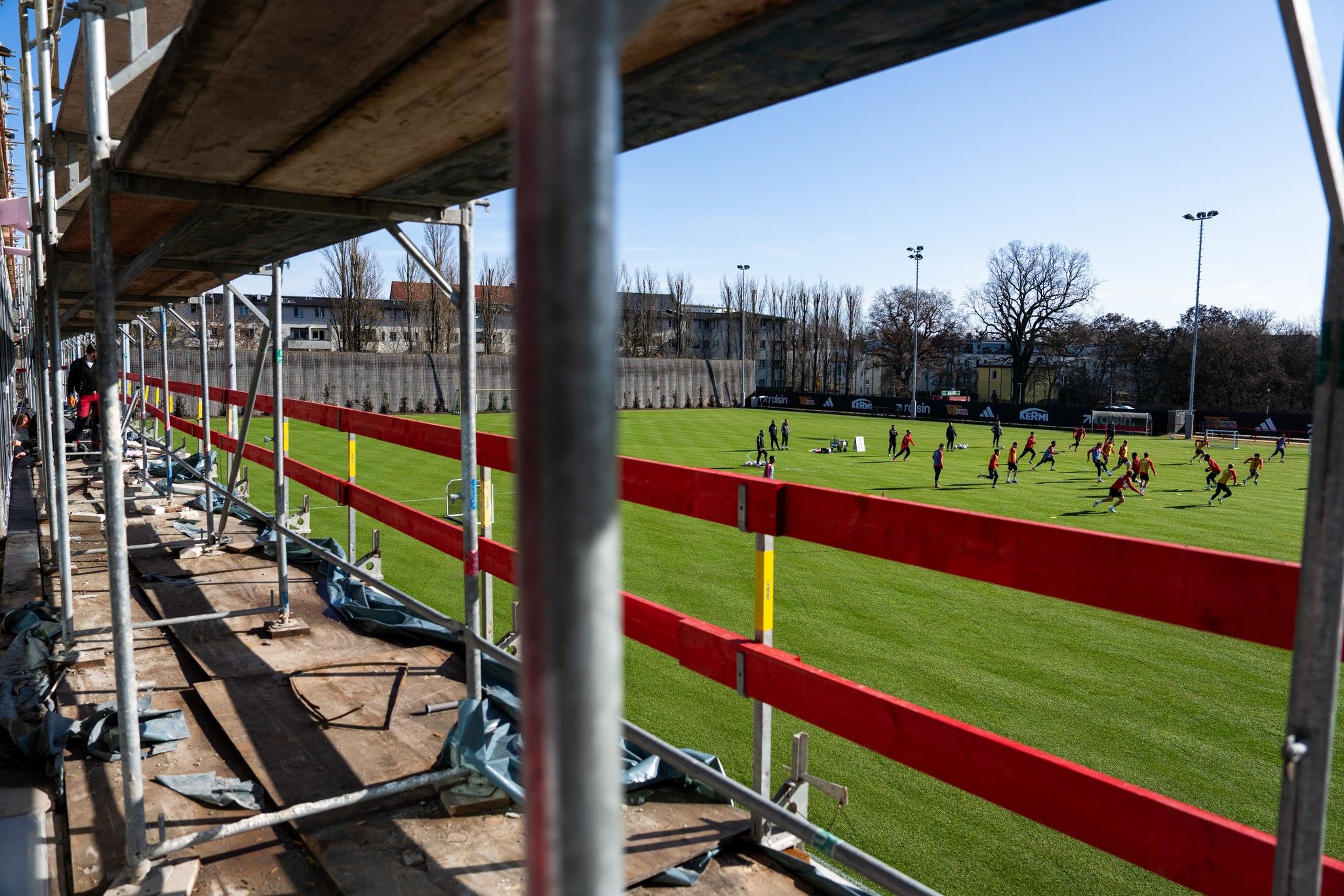 Blick durch ein Gerüst auf ein Fußballfeld, auf dem viele Spieler trainieren, unter klarem Himmel.