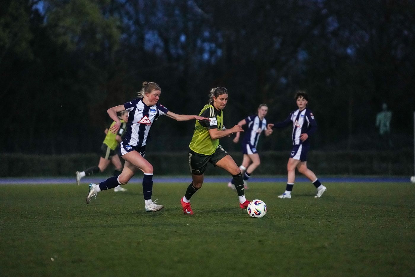 Zwei Fußballspielerinnen kämpfen um den Ball auf einem Spielfeld bei Dämmerung.