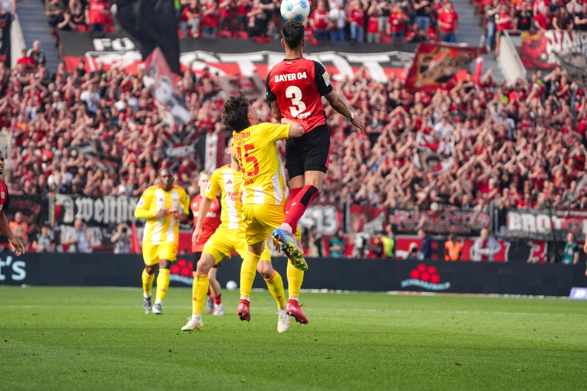 Ein Fußballspieler springt hoch, um den Ball zu erreichen, während ein Gegner ihn verteidigt. Im Hintergrund jubelnde Fans.