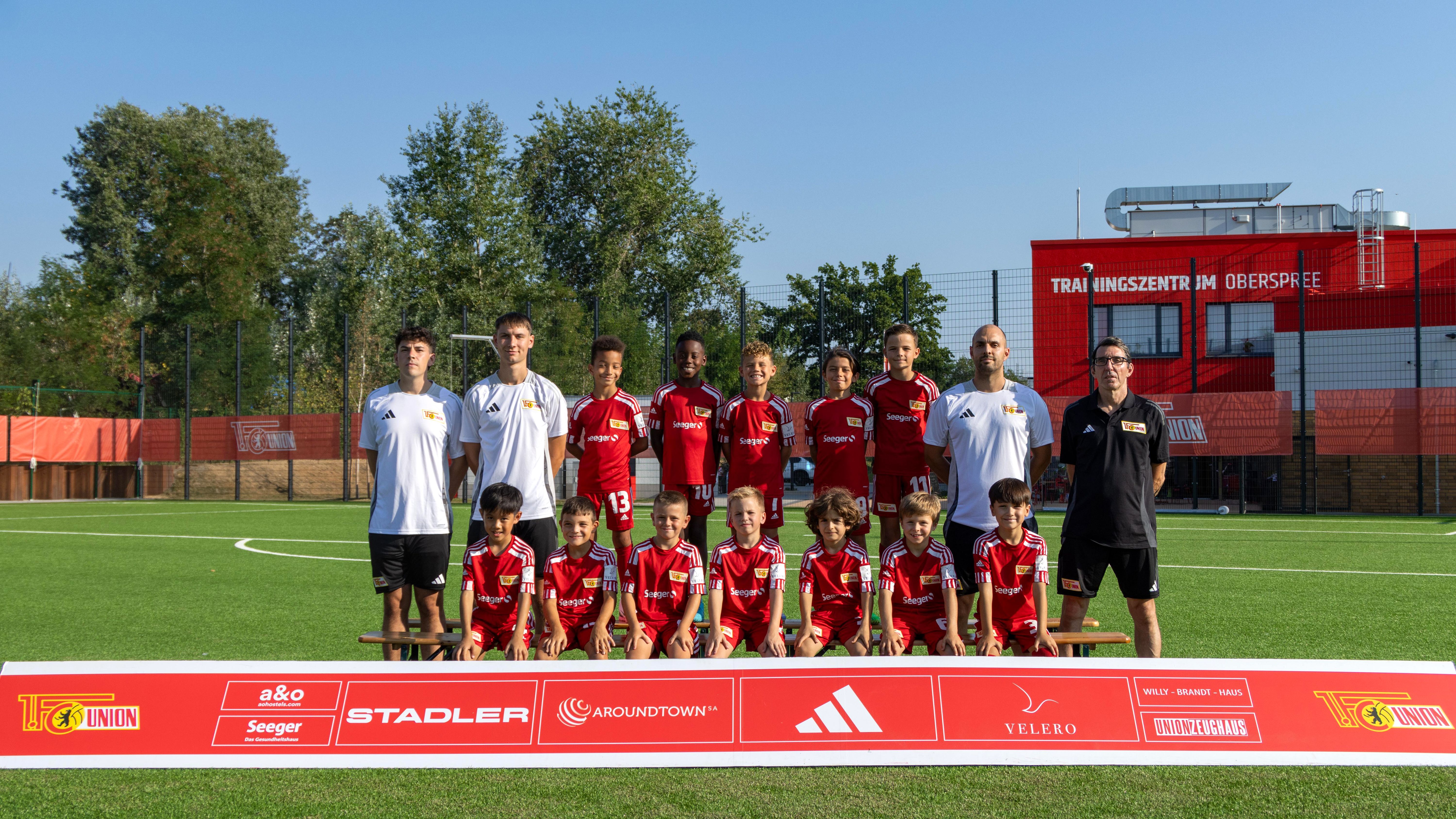 Eine Fußballmannschaft steht auf einem Platz, umgeben von Trainern, mit Trikots in Rot und Schwarz, unter blauem Himmel.