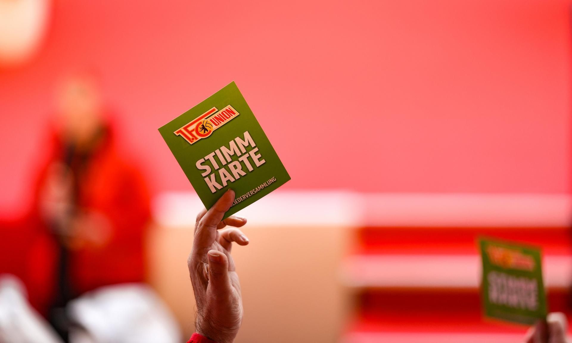 Eine Hand hält eine grüne Stimmkarte mit der Aufschrift "STIMMKARTE" vor einem unscharfen roten Hintergrund.