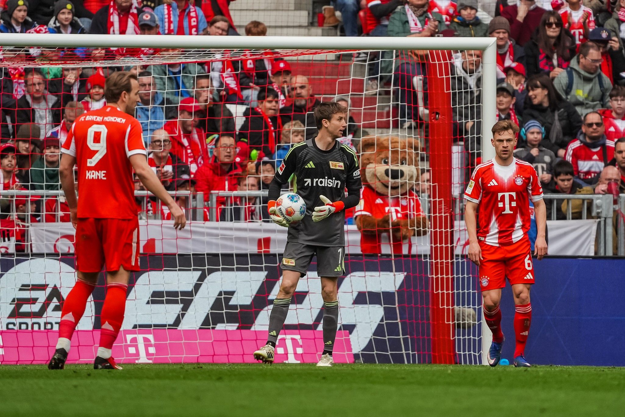 Fußballspiel mit zwei Spielern in roter Uniform, einem Torwart in grauer Ausrüstung und Fans im Hintergrund.