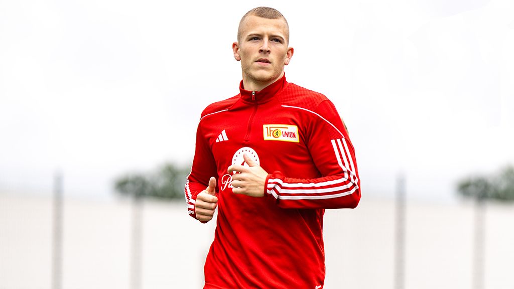 A soccer player in a red tracksuit is jogging on a sports field.