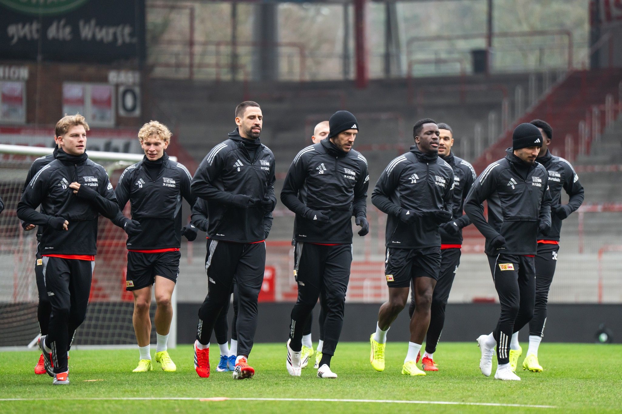 Fußballmannschaft in schwarzen Trainingsanzügen joggt auf dem Platz bei leichtem Regen.