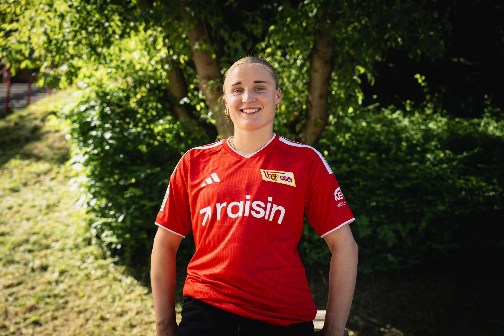 Eine lächelnde Person trägt ein rotes Trikot mit dem Schriftzug „raisin“ und sitzt vor einem grünen Hintergrund.