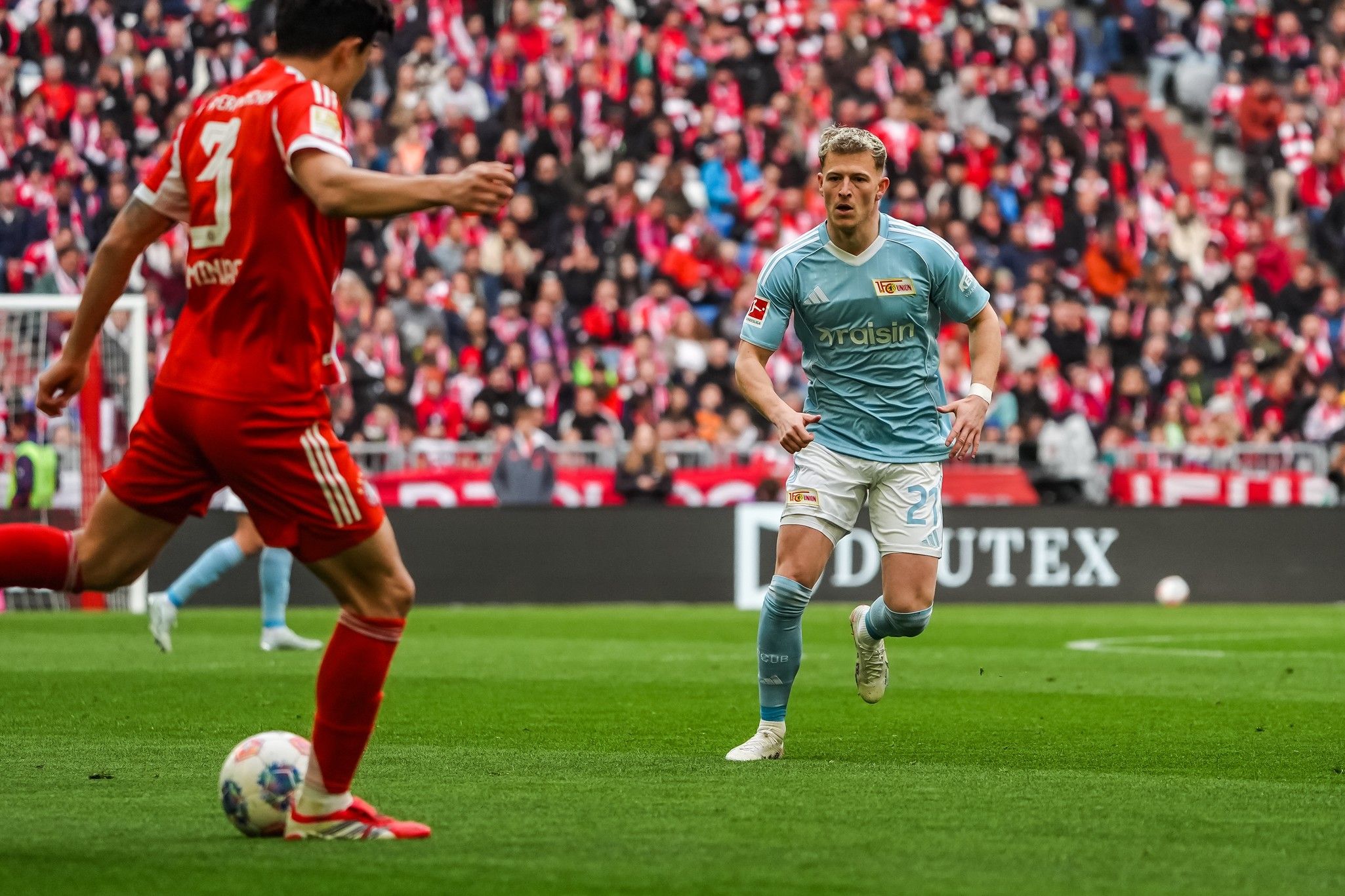 Ein Fußballspieler in blauer Ausrüstung steht auf dem Platz und beobachtet den Spieler mit der Nummer 3 in roter Ausrüstung.