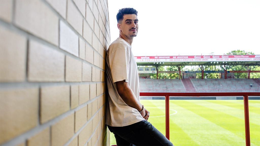 Young man leans against a wall in the stadium, with a view of the field and the stands in the background.