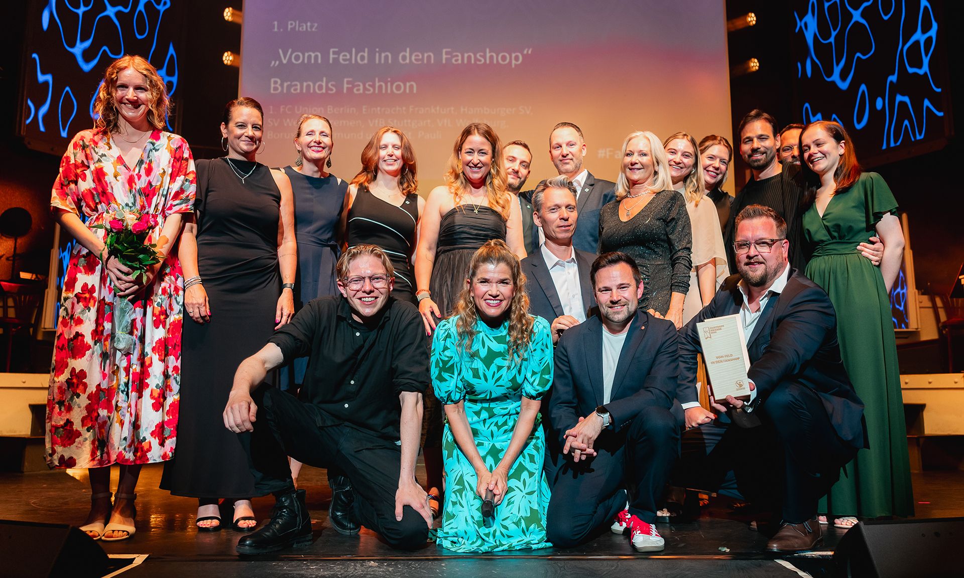Gruppenfoto von Teilnehmern und Gewinnern einer Preisverleihung auf einer Bühne, umgeben von festlicher Beleuchtung.