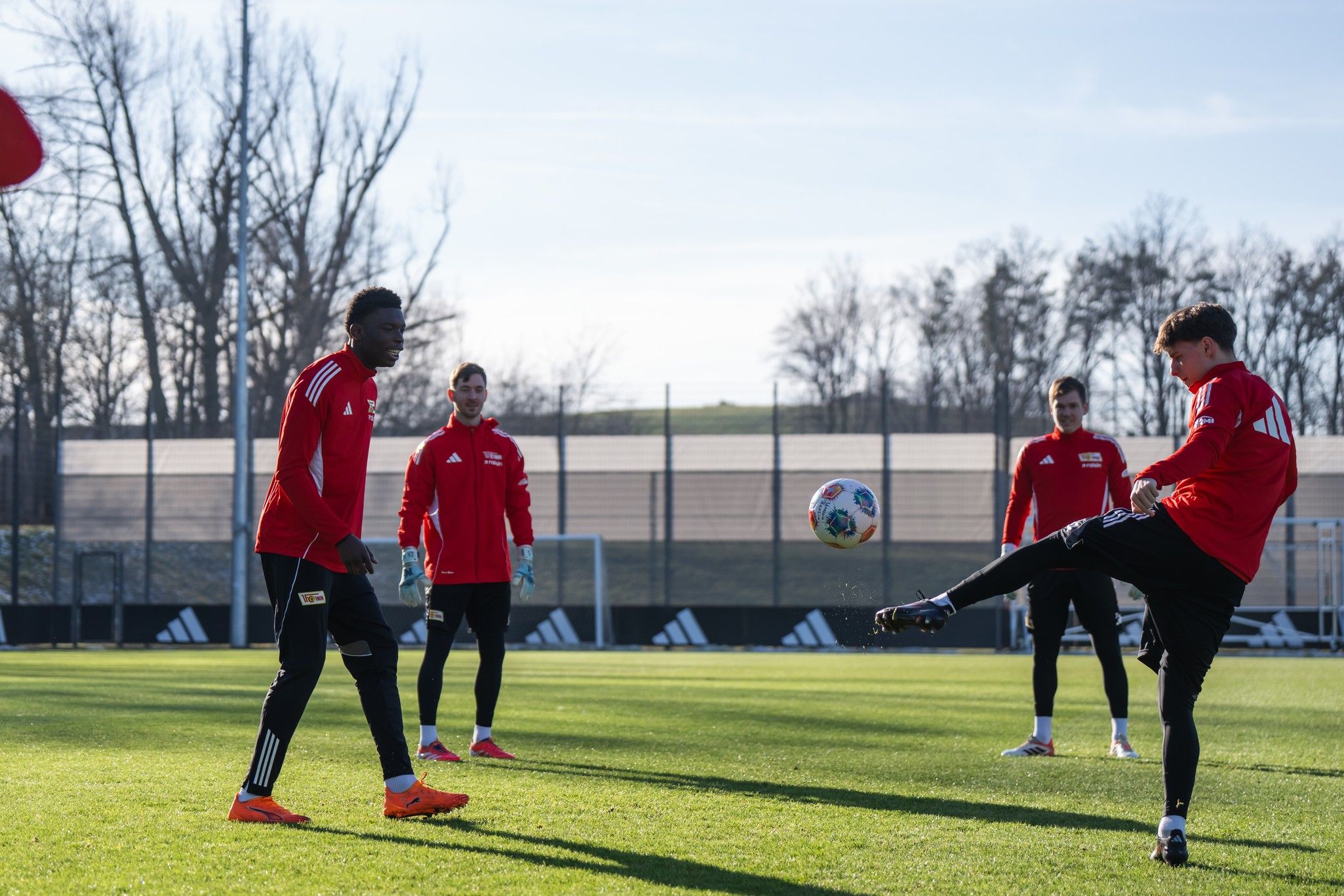 Fünf Fußballspieler in roten Trikots üben auf einem Trainingsplatz unter blauem Himmel und Sonnenlicht.